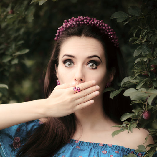 Picture of a lady wearing handmade Boho Chic Jewelry: hazel-colored eyes, wide, beautiful lady: pale skin and brown long hair in a blue off-the-shoulder dress, purple jewelry hair piece, earrings and matching ring, with her hand over her mouth