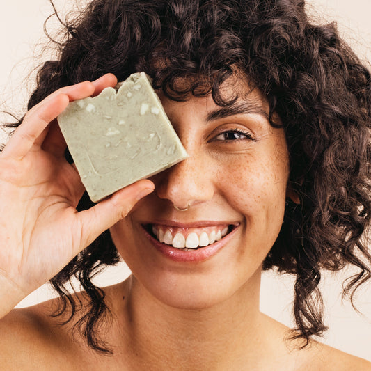 A smiling female soap maker proudly holds up her freshly crafted soap, bringing it close to her face to admire the intricate details and the sweet aroma of her creation.