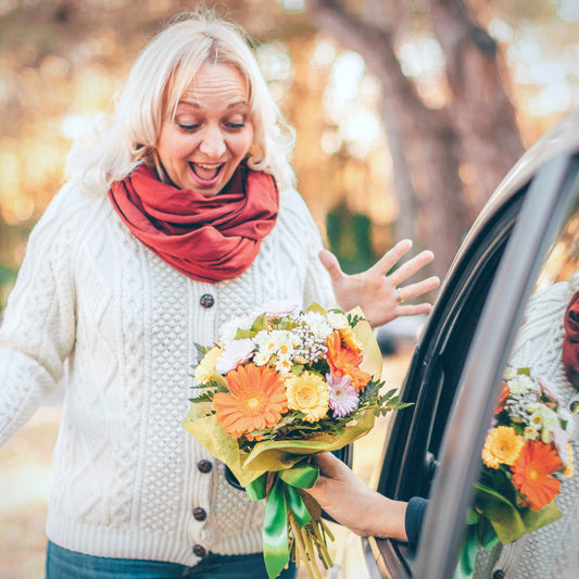 Picture of blond hair excited middle-aged lady looking surprised at the flower bouquet given by her man who sits in a car. Shop local gifts at LocalWe.