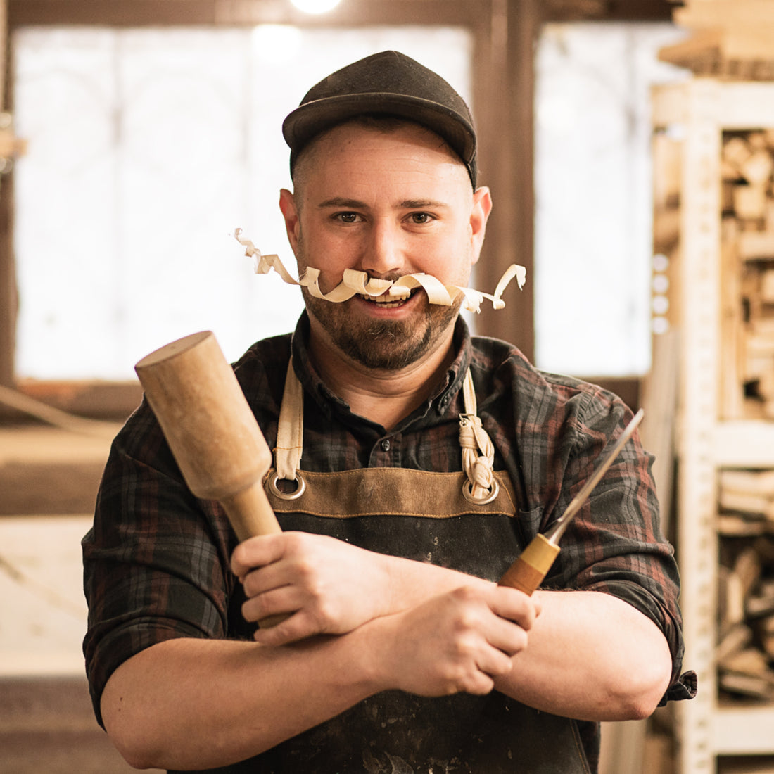 Woodworker with a curly wood shaving mustache and crossed arms holding a mallet and awl