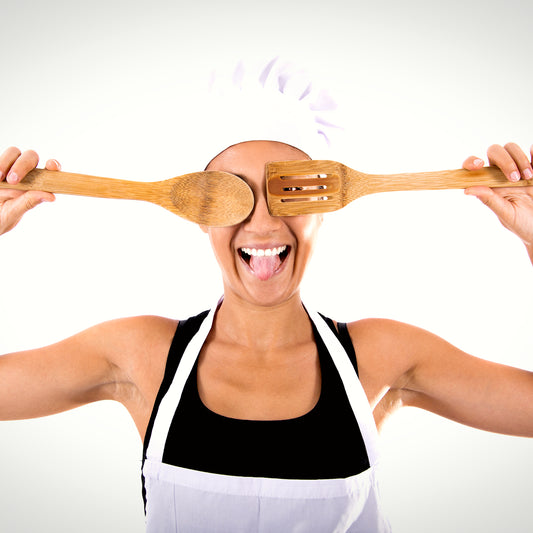 A smiling women in a chef's hat holding over her eyes handmade wooden utensils that are made from urban wood