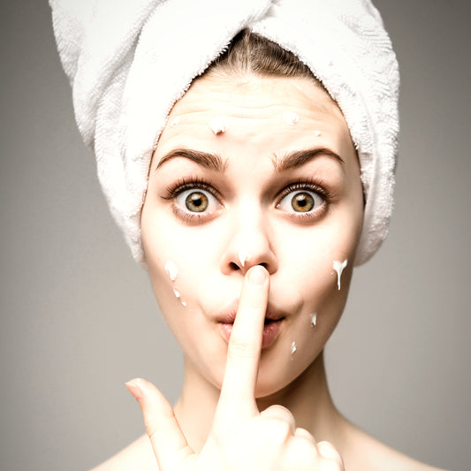 funny pretty young woman with a white towel wrapped around her drying hair, with white skincare cream on her face and her finger on her nose